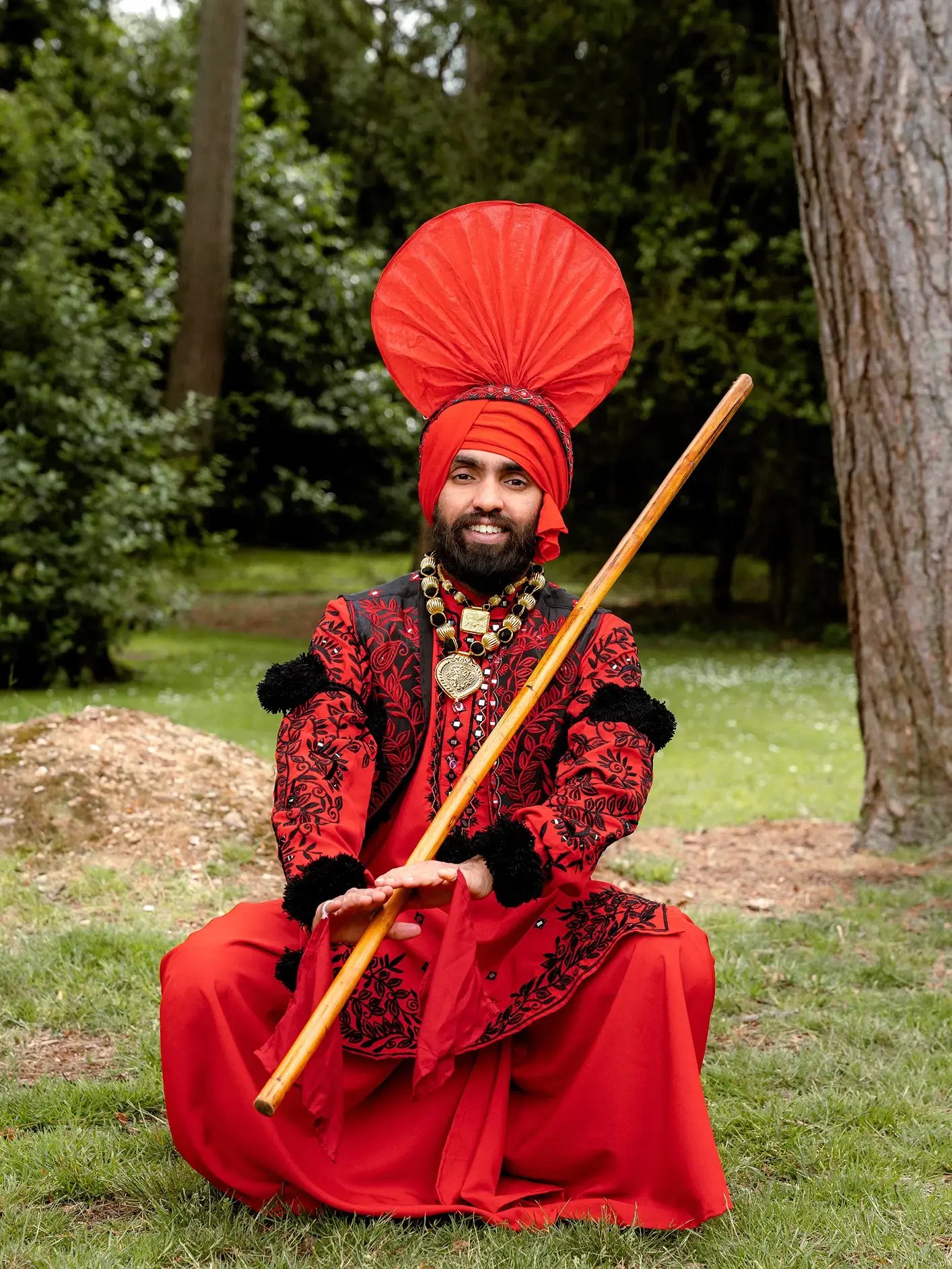 Man wearing traditional red Punjabi Bhangra attire and turban, holding a wooden staff while posing outdoors.