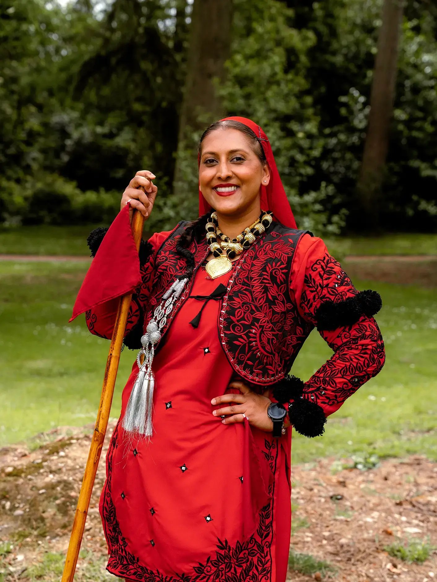 Woman wearing traditional red Punjabi dance attire and jewellery, holding a wooden staff while posing outdoors.