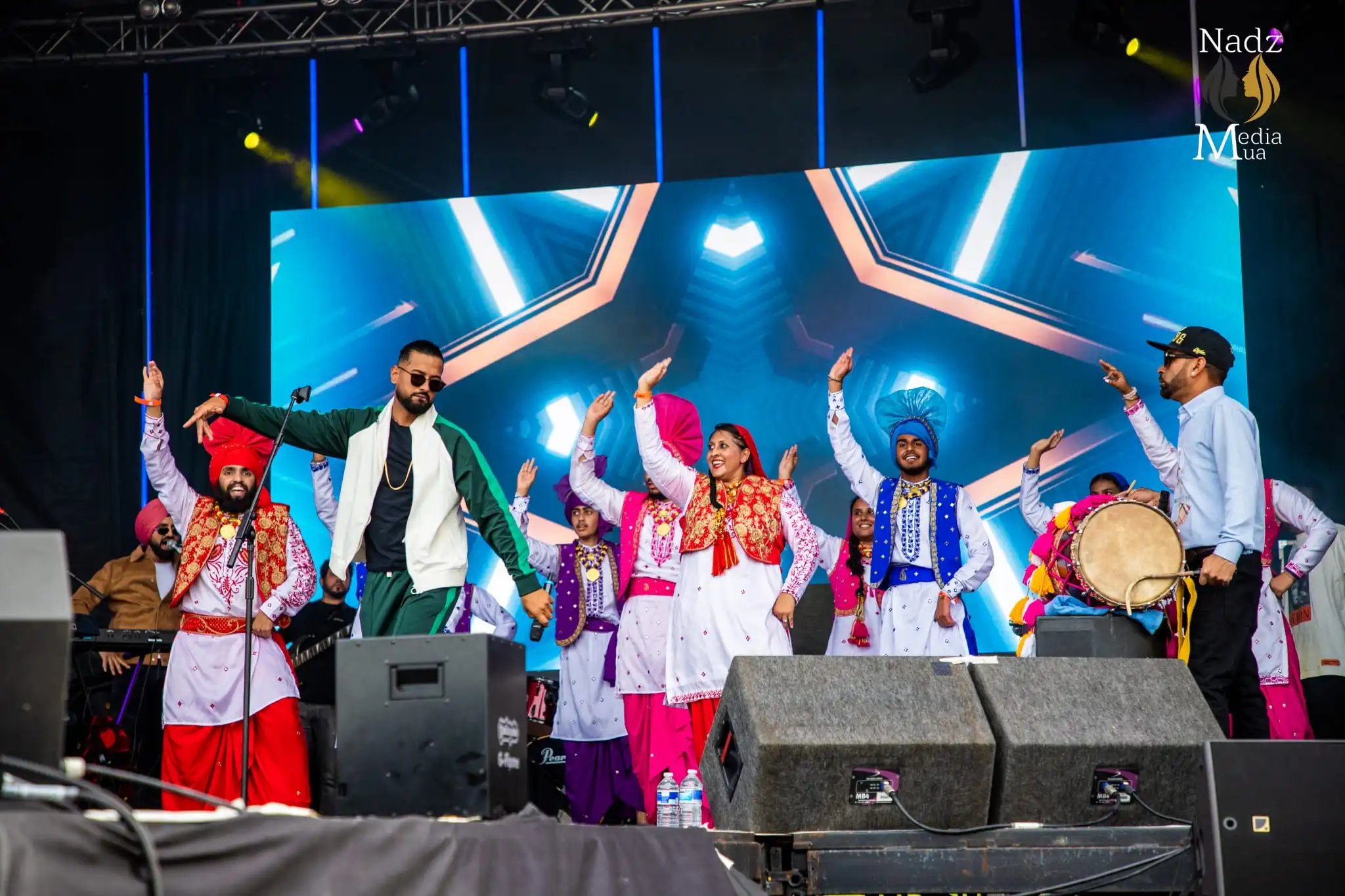 Bhangra dancers in colourful traditional Punjabi costumes performing on stage alongside a male singer and dhol player.