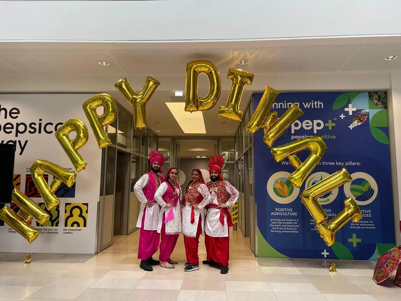 Four Bhangra dancers in traditional Punjabi costumes posing beneath gold Happy Diwali balloons at a corporate event.
