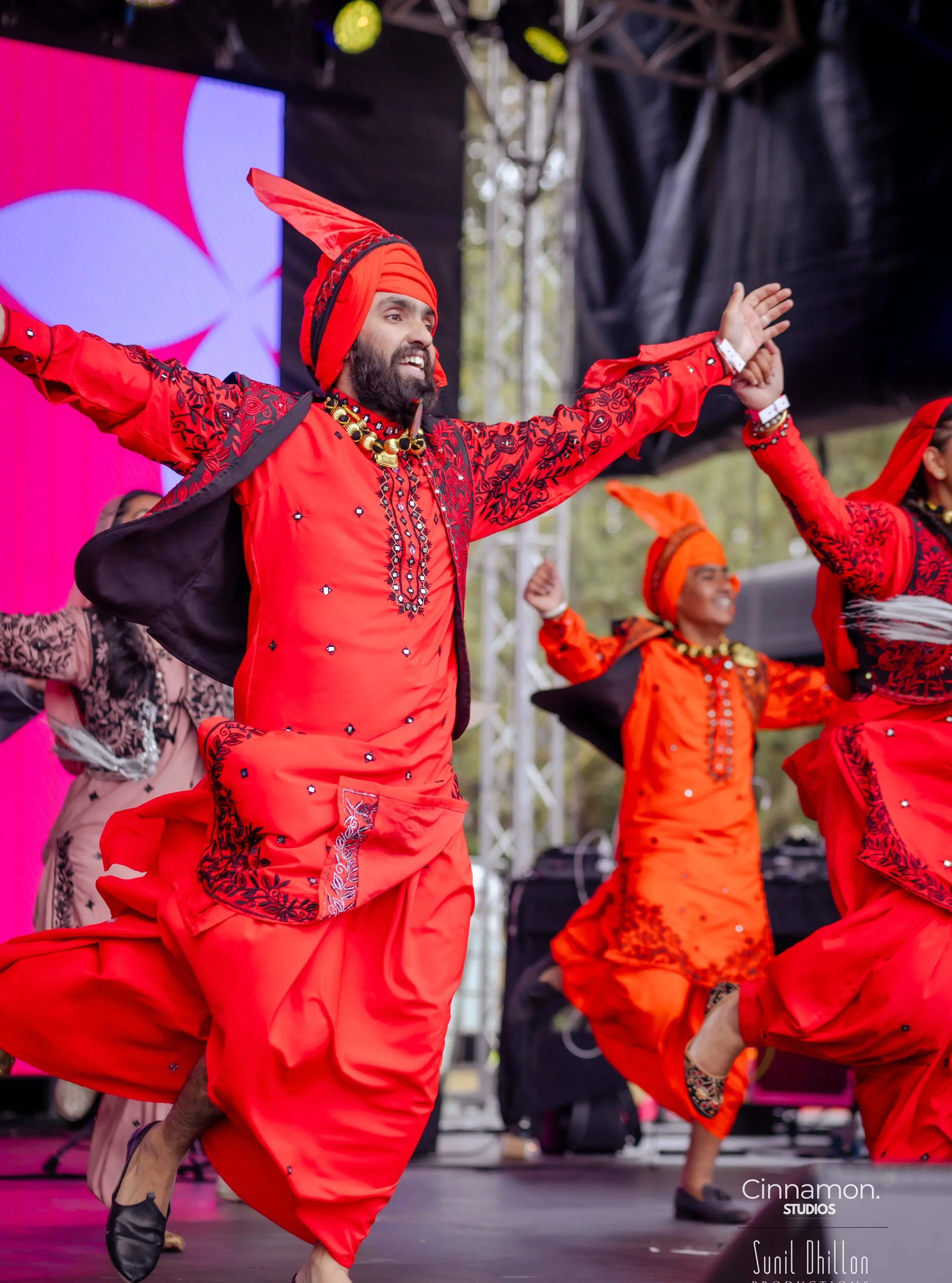 Bhangra dancer performing energetically on stage in a red traditional Punjabi costume with other dancers behind him.