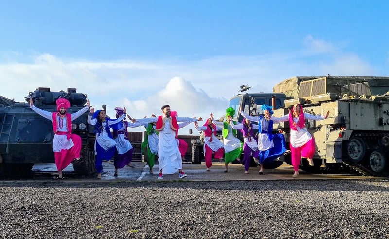 Group of Bhangra dancers in colourful traditional Punjabi costumes performing outdoors between military vehicles.