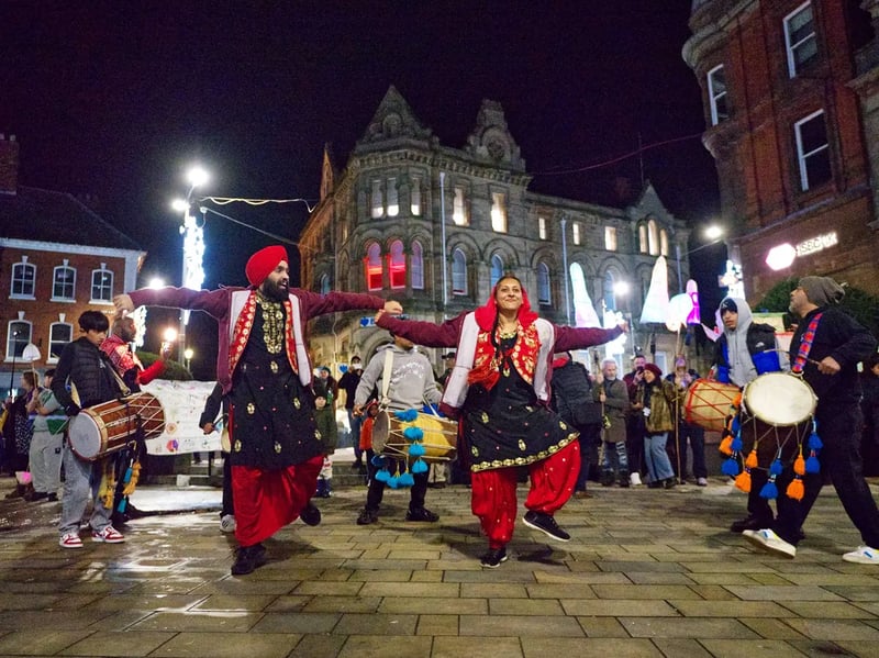 Two Bhangra dancers performing outdoors at night alongside dhol players, with a crowd gathered in a town square.