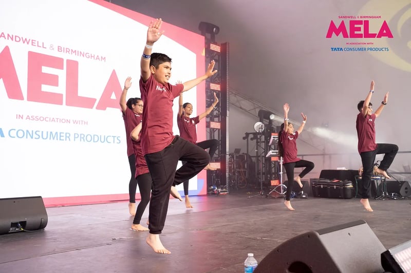 Young Bhangra dancers in matching maroon shirts performing a coordinated routine on stage at Sandwell and Birmingham Mela.