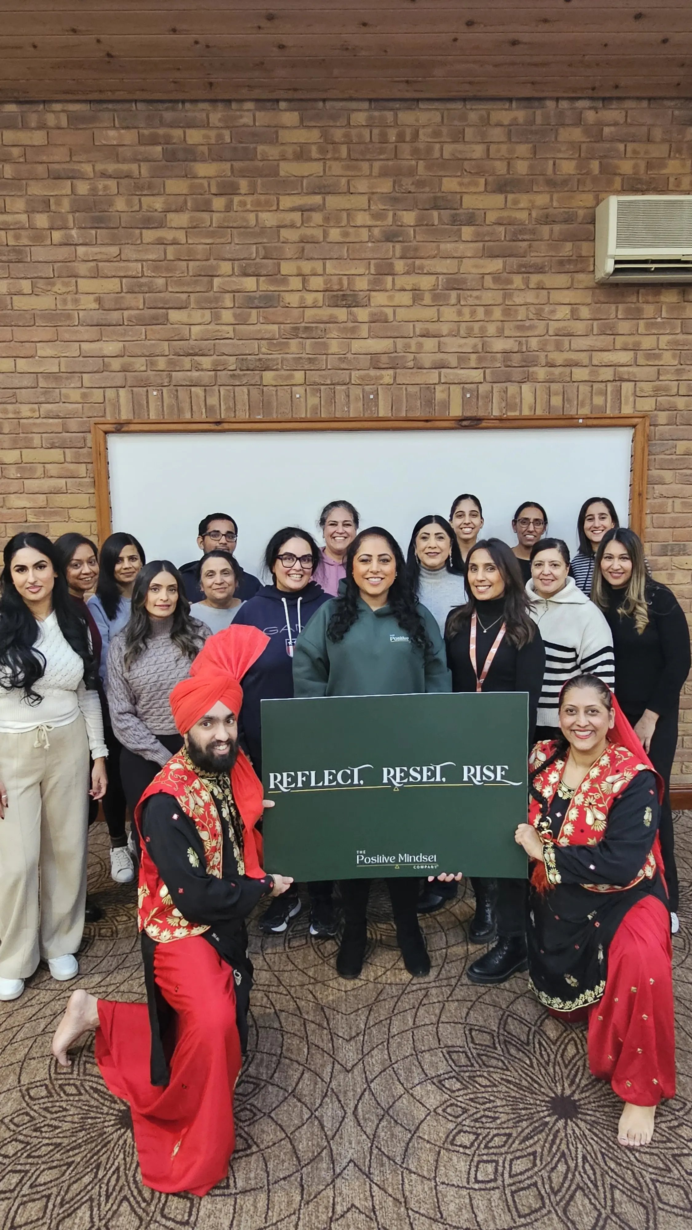 Community group posing together at a wellbeing workshop, with two Bhangra dancers kneeling at the front beside a “Reflect. Reset. Rise.” sign.