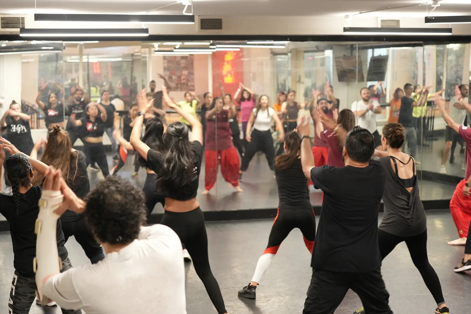 Large group taking part in a Bhangra dance class in a mirrored studio, following a coordinated movement with raised arms.